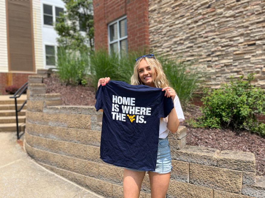 Student standing in front of Vandalia Apartments holding a tee shirt that says "Home Is Where The *flying WV* Is."